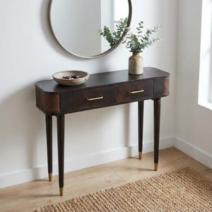 Op de afbeelding: Donkerbruine houten console met twee lades en messing handgrepen. De tafel heeft aan elk uiteinde een geweven rotan paneel en staat op vier taps toelopende poten met goudkleurige uiteinden. Boven de tafel hangt een ronde spiegel.