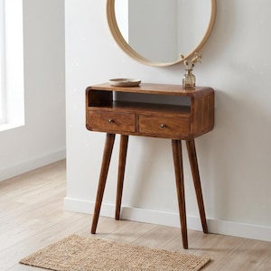 Può includere: Una consolle in legno in stile moderno della metà del secolo, con due cassetti e gambe affusolate. Il tavolo è di colore marrone caldo e ha un piano arrotondato. Uno specchio rotondo è appeso sopra il tavolo e un piccolo tappeto tessuto è sul pavimento.
