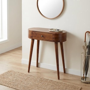 Op de afbeelding: Een middelbruine houten sidetable met vier taps toelopende poten en een enkele centrale lade met een kleine ronde knop. Op het tafelblad staat een ondiep, lichtgekleurd ovaal dienblad en aan de muur erboven hangt een ronde spiegel.