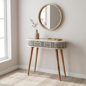 May include: An oval wooden console table with four tapered legs and a drawer featuring a black and white geometric chevron pattern. A round wooden mirror hangs above the table, which holds a beige vase with dried branches and a small wooden tray.