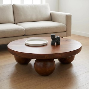 May include: A round, low-profile wooden coffee table with a smooth, dark brown finish, supported by three large, spherical ball-shaped legs. On the tabletop sits a shallow, circular white tray and a small, abstract black stone sculpture.