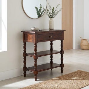 Op de afbeelding: Een donkerbruine houten consoletafel met een lade en twee planken. De tafel heeft gedraaide poten en een kleine messing knop op de lade. Een ronde spiegel hangt aan de muur boven de tafel. Een vaas met groen staat op de tafel.