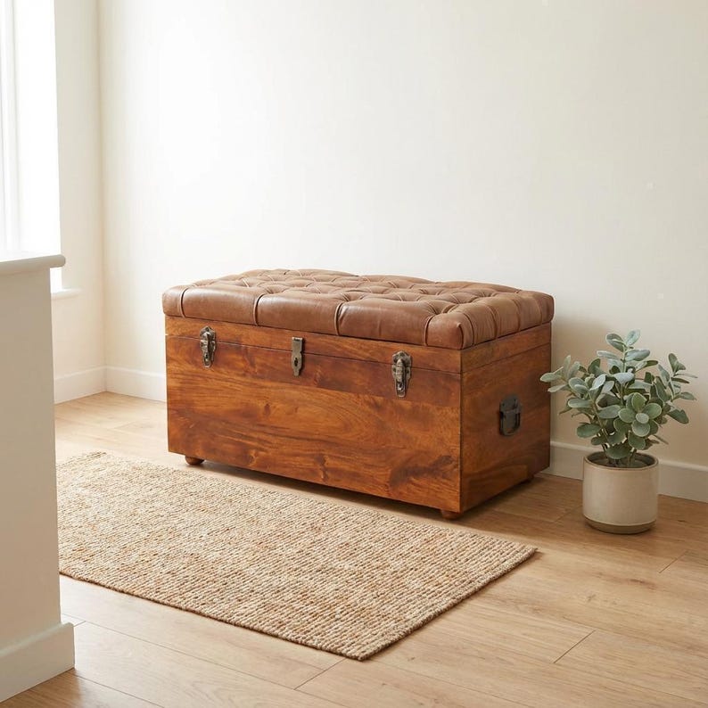 May include: A wooden storage chest with a tufted brown leather top. The chest has metal hardware and is made of a warm-toned wood. A small rug and potted plant are in the foreground.