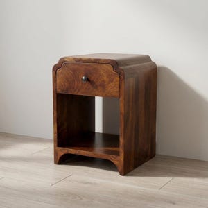 May include: A dark brown wooden bedside table with a single drawer and an open shelf. The table has a curved top and a small, round knob on the drawer. The wood grain is visible, and the table is approximately 61 cm tall.
