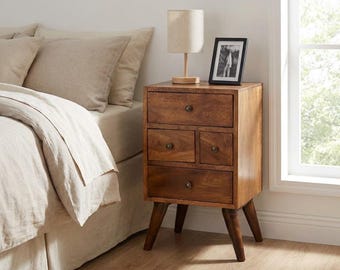 May include: A wooden bedside table with four drawers, featuring a top drawer, two smaller side-by-side drawers, and a bottom drawer, all with round knobs. The table stands on four tapered legs and holds a small lamp and a framed photograph.