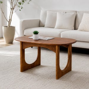 Op de afbeelding: Een ovale houten salontafel met een warme bruine afwerking, voorzien van twee gebogen zijsteunen. Een kleine witte keramische pot met een groene vetplant en een dun wit boek liggen op het gladde, vlakke oppervlak van het tafelblad.