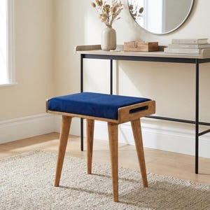 May include: A wooden vanity stool with a rectangular, navy blue velvet seat. The stool has four angled, light brown wooden legs. The frame of the seat is also light brown wood, with a cutout on each side. The stool is on a beige rug.