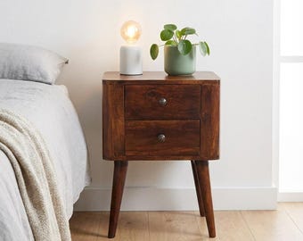 May include: A dark brown wooden bedside table with two drawers, each featuring a small round metal knob. The table stands on four tapered legs and holds a white cylindrical lamp with a glowing bulb and a green potted plant in a ceramic pot.