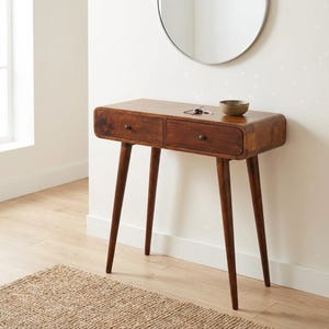 Könnte beinhalten: Eine Konsole im Mid-Century-Modern-Stil aus Holz mit zwei Schubladen und konischen Beinen. Der Tisch hat eine warme braune Farbe und eine abgerundete Front. Eine kleine Schale und Schlüssel liegen obenauf. Der Tisch ist ca. 76 cm lang.