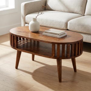 Könnte beinhalten: Ovaler Couchtisch aus Holz mit einem unteren Regal und Lamellen an den Seiten. Der Tisch hat vier konische Beine und ist in einem warmen Braunton gebeizt. Eine weiße Vase mit Zweig und zwei Büchern ruht auf der Tischplatte.