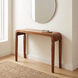 May include: A wooden console table, approximately 120 cm long, with a curved top and four slender legs. The table is a warm brown colour and has a small tray on top. A round mirror hangs on the wall.