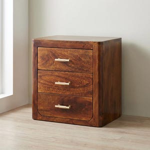 Puede incluir: Una mesita de noche de madera con tres cajones y un acabado marrón cálido. La mesita tiene bordes redondeados y tiradores dorados. La veta de la madera es visible en los frentes y laterales de los cajones. Medidas en cm.