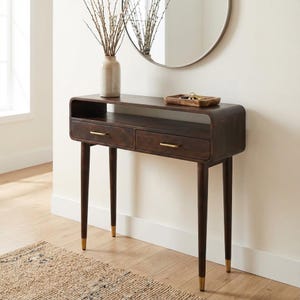 Op de afbeelding: Een donkerbruine houten console met twee lades en messing hardware. De tafel heeft een gebogen bovenkant en slanke poten met messing uiteinden. Een vaas met takken en een ronde spiegel zijn zichtbaar.
