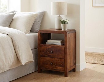 Könnte beinhalten: Ein dunkelbrauner Nachttisch aus Holz mit zwei Schubladen und einem offenen Fach. Der Nachttisch hat abgerundete Ecken und kleine runde Knöpfe. Eine beige Lampe und eine kleine grüne Topfpflanze stehen oben, zwei Bücher liegen im offenen Fach.