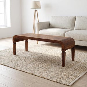 May include: A dark brown wooden coffee table with a curved top and four sturdy legs. The table sits on a beige woven rug in a living room setting. A light-colored sofa and a floor lamp are in the background.