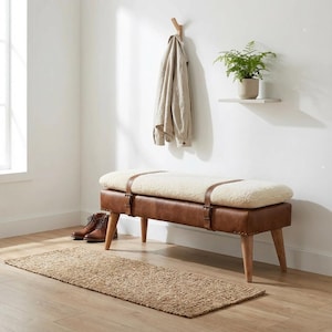 May include: A rectangular bench featuring a brown leather base with four wooden legs, topped with a thick cream-coloured sherpa cushion secured by two brown leather straps. The bench is positioned on a rectangular woven jute rug.