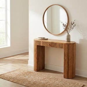 May include: A wooden console table with a round mirror above it. The table has a ribbed design and a small drawer. A small vase with dried flowers sits on the table. A woven rug is on the floor.