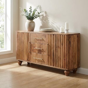 May include: A wooden sideboard featuring three central drawers with thin gold-toned horizontal handles and two side cabinets with vertical fluted detailing. The piece stands on four rounded feet and is topped with a ceramic vase of greenery and two white candles.