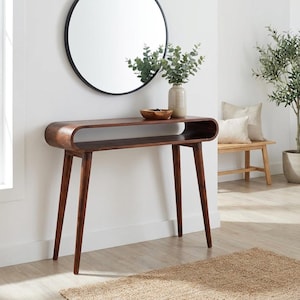May include: A mid-century modern style, dark brown wooden console table with a curved top and lower shelf. The table has four angled legs. A round black mirror hangs above the table, which is decorated with a vase of greenery and a wooden bowl.