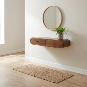 May include: A wall-mounted floating console table made of medium-toned wood with two drawers and rounded edges. Above the console is a circular mirror with a light wood frame. A small green fern in a gray pot sits on the console surface.