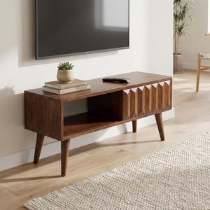 May include: A dark brown wooden TV stand in a mid-century modern style. The unit has a rectangular shape with angled legs and a geometric design on the right. It includes an open shelf and a closed compartment.