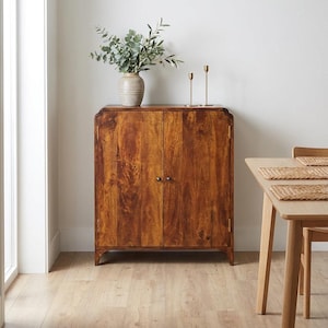 Puede incluir: Un mueble de madera de dos puertas con la parte superior curvada, en un tono marrón cálido con veta de madera visible. Un jarrón con vegetación y dos candelabros dorados encima. El mueble está en una habitación con suelo de madera y una mesa.