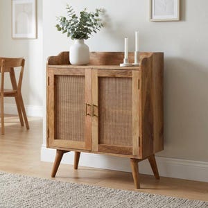 May include: A light brown wooden cabinet with two doors featuring woven cane panels and vertical gold metal handles. The cabinet stands on four tapered legs and holds a white ceramic vase with green foliage and two white pillar candles.