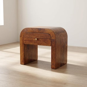 Könnte beinhalten: Ein Nachttisch aus Massivholz mit geschwungener Oberseite und einer Schublade. Der Nachttisch hat eine warme braune Farbe und einen Messingknopf an der Schublade. Das Design ist modern und minimalistisch, etwa 60 cm hoch.