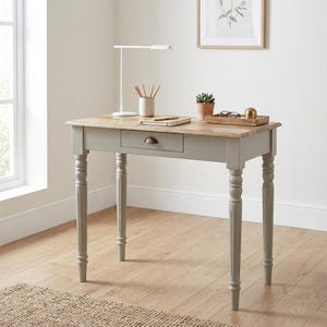 Könnte beinhalten: Ein hellgrauer Schreibtisch aus Holz mit einer naturfarbenen Tischplatte und einer einzelnen Schublade mit kleinem Metallgriff. Der Schreibtisch hat vier gedrechselte Beine und ist mit einer weißen Schreibtischlampe, einem Stiftehalter und einer Pflanze ausgestattet.