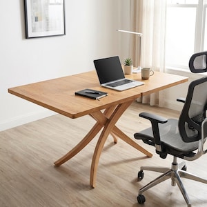 May include: A light brown wooden desk with a laptop, notebook, and mug on top. The desk has a unique cross-legged base and is paired with a black and gray office chair. A small plant and desk lamp are also visible.