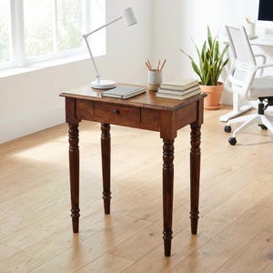 May include: A rectangular wooden desk with a dark brown finish, featuring four turned legs and a single central drawer with a small round knob. The desk surface is smooth and flat, providing a stable area for work or writing tasks.