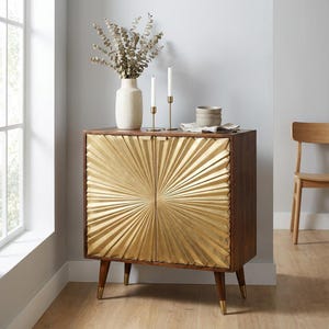 Op de afbeelding: Een houten kast met een gouden zonnestraalontwerp op de deuren. De kast heeft een donkerbruin frame en taps toelopende poten met gouden uiteinden. Een vaas met gedroogde bloemen, kaarsen en kommen staan erop.