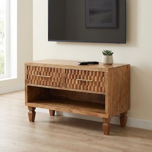 May include: A light brown wooden TV stand with two drawers featuring a geometric pattern and gold-colored handles. The stand has an open shelf below the drawers and sits on four short, carved legs. A remote and a small potted plant are on top.