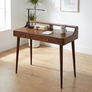 Op de afbeelding: Een donkerbruine houten bureau in mid-century modern stijl. Het heeft twee lades met messing knoppen en taps toelopende poten. Een zwarte bureaulamp, boeken en een mok staan op het bureau. Het bureau is ongeveer 120 cm breed.