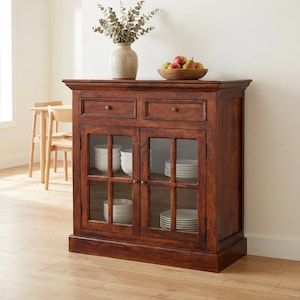 May include: A dark brown wooden cabinet with two drawers and glass-paned doors. Inside the cabinet are white dishes. A vase with dried branches and a wooden bowl of fruit sit on top. The cabinet is in a well-lit room.