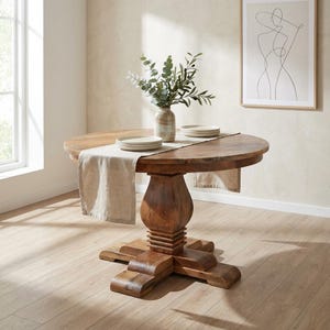 May include: A round wooden dining table with a pedestal base. The table is made of a dark wood and has a natural finish. A beige table runner and plates are on the table. A vase with greenery sits on the table.