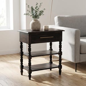 May include: A dark brown wooden side table featuring a single drawer with a thin horizontal metal handle, two lower open shelves, and four turned legs. The table surface holds a textured beige vase with green eucalyptus stems and a small candle.