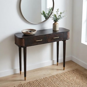 Op de afbeelding: Een donkerbruine houten console met twee lades en messing handgrepen. De tafel heeft geweven zijpanelen en slanke poten met goudkleurige uiteinden. Boven de tafel hangt een ronde spiegel en er staat een vaas met groen.