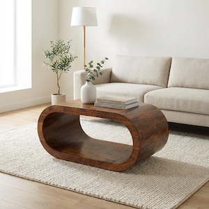 May include: A solid, oval-shaped wooden coffee table with a rich, dark brown finish. The table has a hollowed-out center and is placed on a neutral-colored rug. A beige sofa and a floor lamp are in the background, creating a cozy living room setting.
