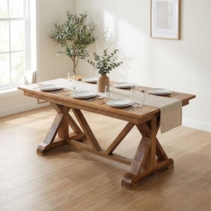 May include: A rectangular wooden dining table with a trestle base, featuring a light beige fabric table runner, six white ceramic plates, six clear glass tumblers, and a central ceramic vase containing green eucalyptus branches.