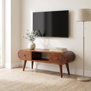 Peut inclure: Meuble TV de style moderne du milieu du siècle en bois brun chaud. Il comporte deux tiroirs, une étagère ouverte et des pieds effilés. Un vase avec de la verdure et des livres sont posés dessus. Une grande télévision noire est fixée au mur.