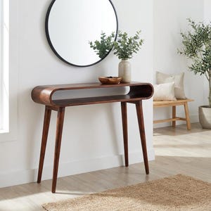 Könnte beinhalten: Dunkelbrauner Konsolentisch im Mid-Century-Modern-Stil mit geschwungener Oberseite und unterem Regal. Der Tisch hat vier konische Beine. Ein runder schwarzer Spiegel hängt darüber. Eine Vase mit Grün und eine Holzschale stehen oben.