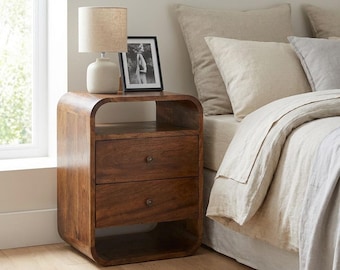 May include: A dark brown wooden bedside table with rounded corners, featuring an open top shelf, two drawers with small round knobs, and an open bottom shelf. A beige table lamp and a black framed photograph sit on the top surface of the bedside table.