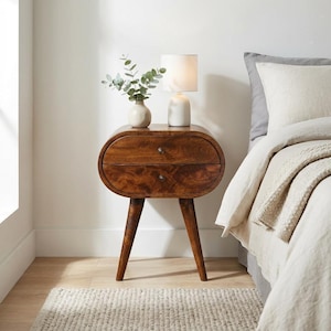 Peut inclure: Une table de chevet en bois avec deux tiroirs et un design incurvé. La table a une finition marron chaud et est soutenue par trois pieds inclinés. Une petite lampe blanche et un vase avec de la verdure sont posés dessus. Un tapis est devant la table.