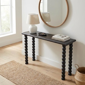 Può includere: Una consolle in legno scuro con gambe tornite, sormontata da una lampada bianca, un piccolo vassoio nero e libri. Uno specchio rotondo è appeso alla parete sopra il tavolo. Un tappeto intrecciato e un cesto sono sul pavimento.