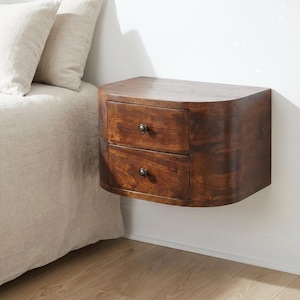 May include: A wall-mounted floating bedside table crafted from dark brown wood with a smooth, curved exterior. This piece features two stacked drawers, each equipped with a single small, dark metal knob centred on the front panel for access.