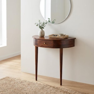 May include: A semi-circular wooden console table with a single drawer featuring a small brass knob. The table stands on three tapered legs. On top sits a light-colored ceramic vase with green foliage and a rectangular woven tray holding a small bowl.