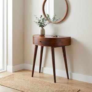 May include: A dark brown, semi-circular console table with a single drawer and four tapered legs. A small vase with greenery and a round mirror are mounted on the wall above the table. The table is made of wood.