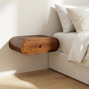 Könnte beinhalten: Ein schwebender Nachttisch aus dunklem Holz mit einer Schublade und einem Messingknopf. Der Nachttisch hat ein abgerundetes, stromlinienförmiges Design und ist an einer weißen Wand befestigt. Die Schlafzimmermöbel stehen neben einem Bett mit weißer Bettwäsche.