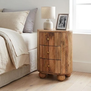 May include: A wooden bedside table with three drawers and a ribbed design. The table has a round shape and sits on four small, rounded legs. A small lamp and a framed picture are on top. The table is next to a bed with beige bedding.
